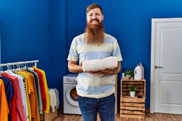 Nuori punapää mies hymyilee luottavainen tilalla taitettu vaatteita pesutupa - Valokuva, kuva