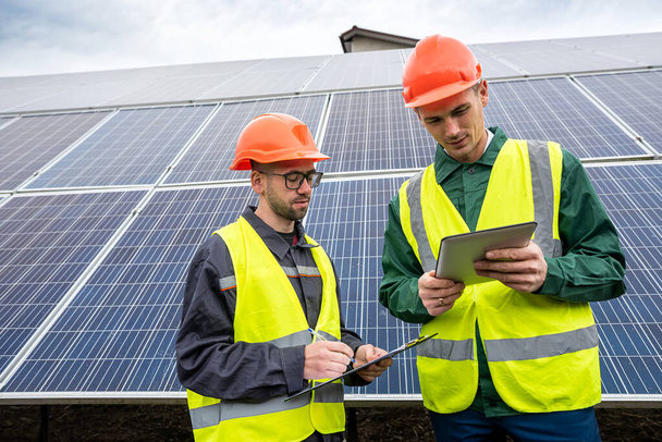 dois jovens homens fortes em capacetes especiais fazem anotações depois de inspecionar os painéis solares. O conceito de economia de energia verde - Foto, Imagem