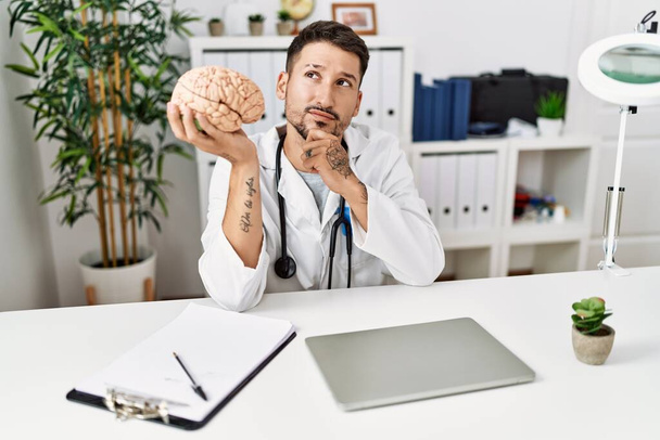 Młody lekarz trzymający mózg w klinice medycznej poważnie myśli o pytaniu z ręką na podbródku, myśląc o mylącym pomyśle  - Zdjęcie, obraz