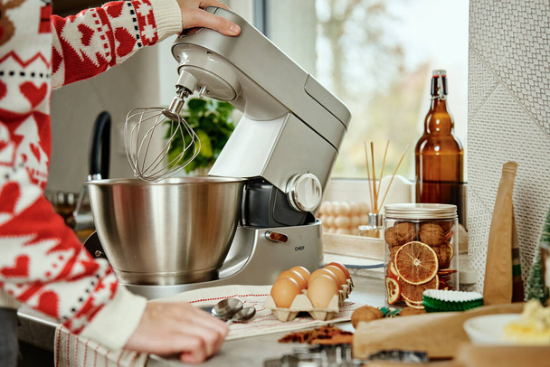 Mujer en jersey navideño festivo preparando masa para galletas en la cocina casera. Las manos femeninas usan mezclador eléctrico. Electrodomésticos de cocina moderna - Foto, Imagen