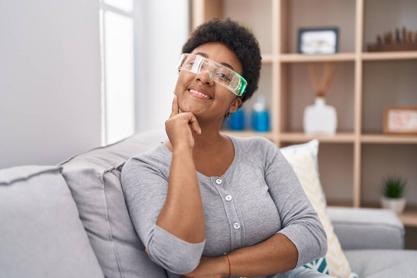 Jeune femme afro-américaine portant des lunettes de réalité virtuelle assise sur le canapé souriant regardant avec confiance la caméra avec les bras croisés et la main sur le menton. penser positivement.  - Photo, image