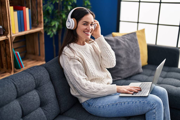 Young woman using laptop and headphones sitting on sofa at home - Фото, изображение