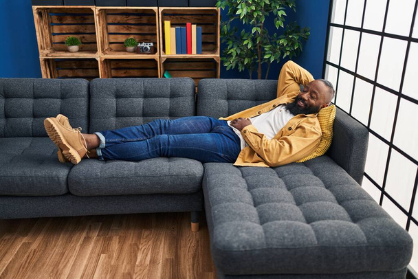 Молодой африканский американец улыбается уверенно расслабленной лежа на диване дома - Фото, изображение