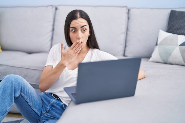 Giovane donna bruna che utilizza il computer portatile a casa che copre la bocca con la mano, scioccata e spaventata per errore. espressione sorpresa  - Foto, immagini