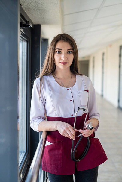 Μια νοσοκόμα με μια ελκυστική εμφάνιση σε μια στολή περιμένει τους ασθενείς στην κλινική στο διάδρομο. Η έννοια του γυναικείου πορτρέτου. Γιατρός με στολή. - Φωτογραφία, εικόνα