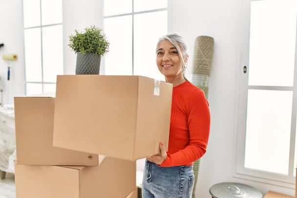 Mujer de pelo gris de mediana edad sonriendo confiado sosteniendo paquete en el nuevo hogar - Foto, Imagen