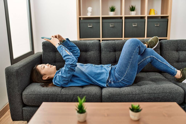 Junge Chinesin mit Smartphone zu Hause auf dem Sofa liegend. - Foto, Bild