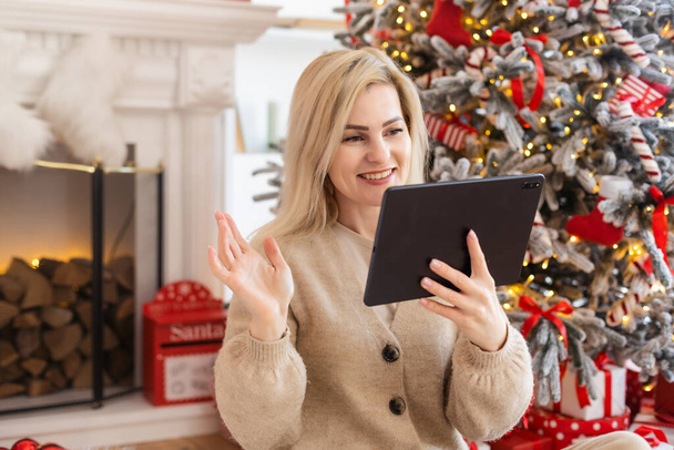 Christmas online holiday. Happy woman having video call with their family or friends. Young woman uses a digital tablet near decorated festive tree at home. Virtual meeting - Foto, Imagem