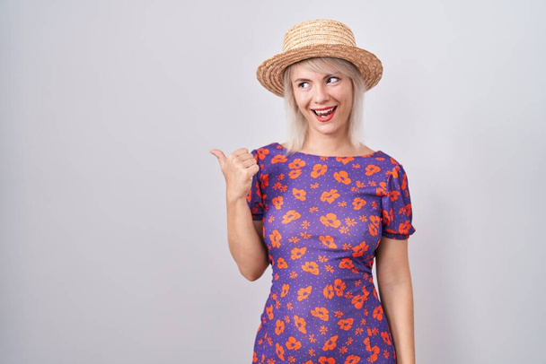 Fiatal kaukázusi nő virágos ruhában és nyári kalapban mosolyog boldog arccal néz, és mutatva oldalra hüvelykujjával felfelé.  - Fotó, kép