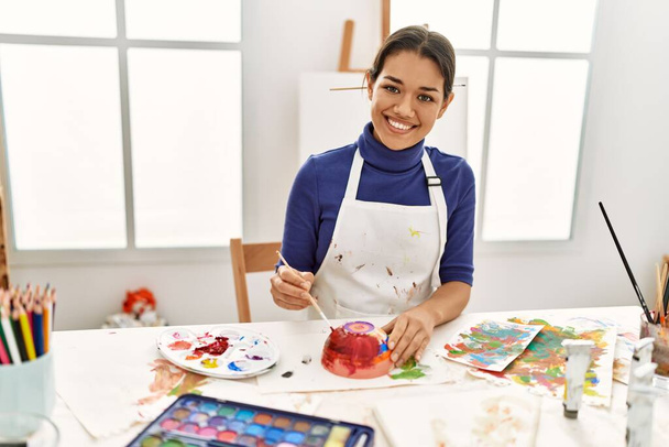 Jovem mulher latina sorrindo confiante pintura cerâmica argila no estúdio de arte - Foto, Imagem