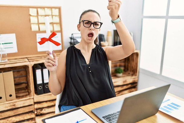 Young brunette woman holding graduate degree diploma at the office annoyed and frustrated shouting with anger, yelling crazy with anger and hand raised  - 写真・画像