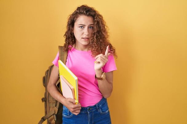 Jovem caucasiana vestindo mochila estudantil e segurando livros apontando para o lado preocupado e nervoso com indicador, expressão preocupada e surpreso  - Foto, Imagem