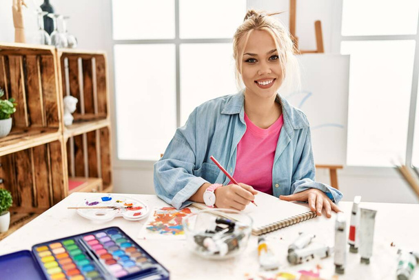 Νεαρή καυκάσια γυναίκα καλλιτέχνης χαμογελώντας αυτοπεποίθηση σχέδιο στο σημειωματάριο τέχνης στο στούντιο - Φωτογραφία, εικόνα