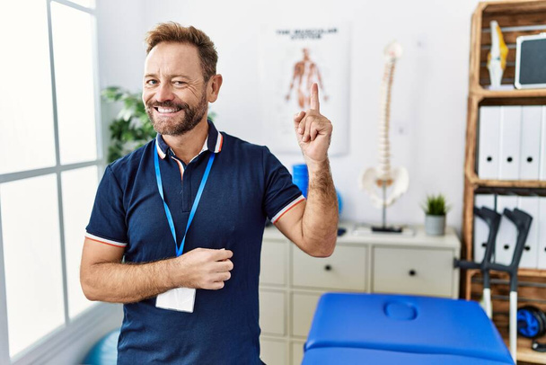 Physiothérapeute d'âge moyen homme travaillant à la clinique de récupération de la douleur avec un grand sourire sur le visage, pointant avec la main et le doigt vers le côté regardant la caméra.  - Photo, image