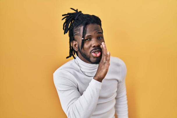 African man with dreadlocks wearing turtleneck sweater over yellow background hand on mouth telling secret rumor, whispering malicious talk conversation  - Zdjęcie, obraz