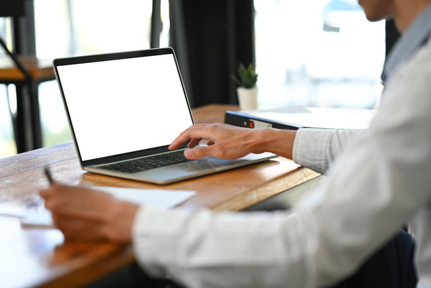 Captura recortada de trabajador de oficina masculino usando computadora portátil y escribiendo información en el bloc de notas. - Foto, Imagen