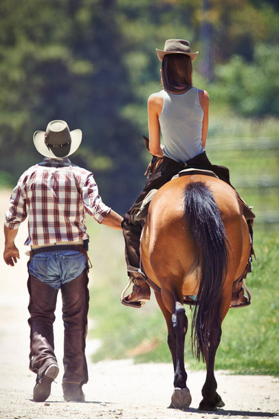 Podoba mi się na świeżym powietrzu. Widok z tyłu kowboja prowadzącego młodą kobietę na koniu wzdłuż wiejskiego pasa - Zdjęcie, obraz