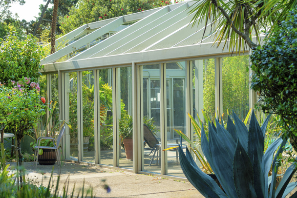 Slunečně osvětlený skleník na zahradě se stromy a listím. Dvoulůžkový skleníkový skleník s dveřmi a posezením se zastíněnými rostlinami v popředí v příměstské oblasti. - Fotografie, Obrázek