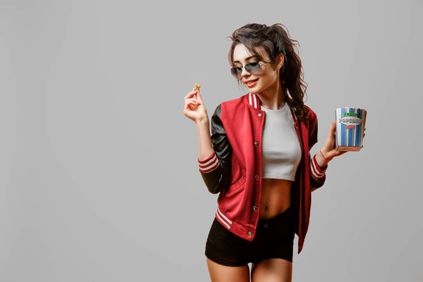 Portrait of pretty stylish brunette girl in sunglasses holding pop corn bowl over gray background. Space for text  - Foto, afbeelding