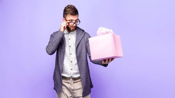 Handsome bearded man looks skeptically at a pink gift box holding it in his hand and adjusting his glasses isolated on purple background  - Fotó, kép