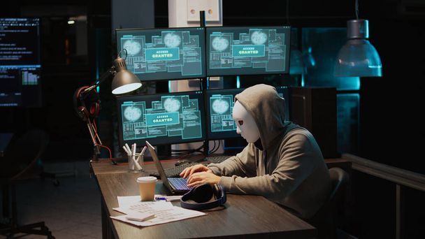 Maskierte Kriminelle hacken nachts Computerserver, arbeiten am Laptop und an mehreren Monitoren, um IT-Informationen zu stehlen. Hacker mit versteckter Identität agieren gefährlich, Cloud Computing. Handschuss. - Foto, Bild