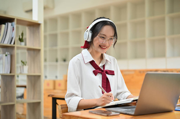 Piękna i mądra młoda studentka Azji w okularach i słuchawkach, studiująca na zajęciach online w bibliotece kampusu. - Zdjęcie, obraz