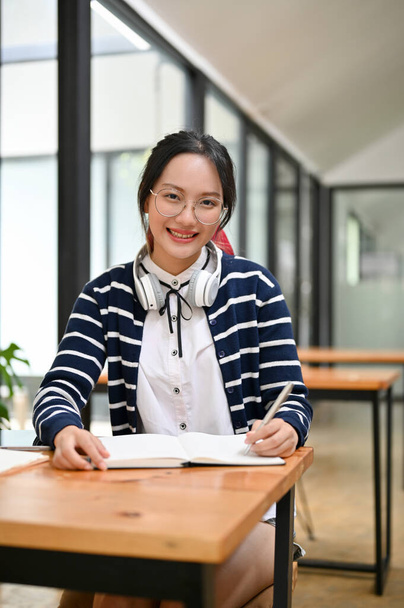 Retrato, Hermosa y encantadora joven asiática con auriculares haciendo la tarea, escribiendo algo en su cuaderno mientras está sentada en el espacio de co-trabajo de la cafetería. - Foto, Imagen