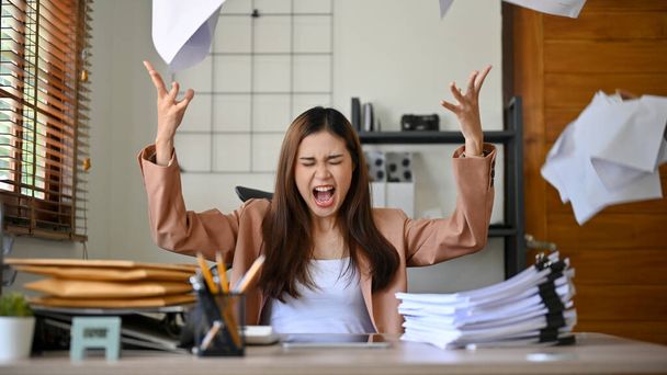 Enfurecida, furiosa, loca y loca empresaria asiática milenaria o trabajadora de oficina gritando, gritando y vomitando papeles en el escritorio de su oficina. Depresión, exceso de trabajo, fracaso. - Foto, Imagen