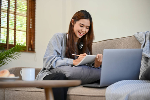 Aantrekkelijke en gelukkige jonge Aziatische vrouwelijke student zit op haar comfortabele bank, het doen van huiswerk, het schrijven van een essay in haar woonkamer.  - Foto, afbeelding