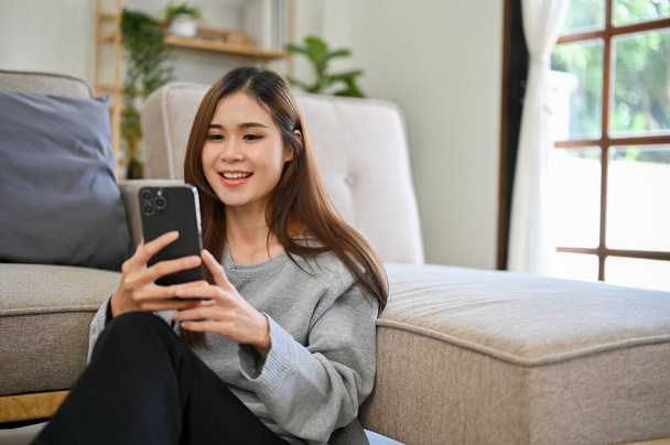 Расслабленная и охлаждающая молодая азиатка в повседневной одежде, сидит в своей удобной гостиной и использует свой смартфон, чтобы общаться со своими друзьями. - Фото, изображение