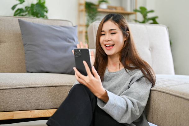 Atrakcyjna młoda Azjatka na swoim smartfonie rozmawiająca lub dzwoniąca wideo z przyjaciółmi relaksując się w wygodnym salonie. - Zdjęcie, obraz