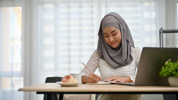 オフィスで働くヒジャーブを身に着けている魅力的で専門的な千年紀アジアのイスラム教徒のビジネス女性,彼女のノートに書き込みやメモを取る. - 写真・画像