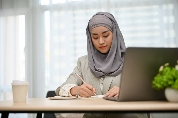 Professionelle und schöne asiatische muslimische Geschäftsfrau oder Geschäftsfrau, die sich auf ihre Projektaufgabe konzentriert und ihren Plan an ihrem Schreibtisch auf ein Buch schreibt. - Foto, Bild