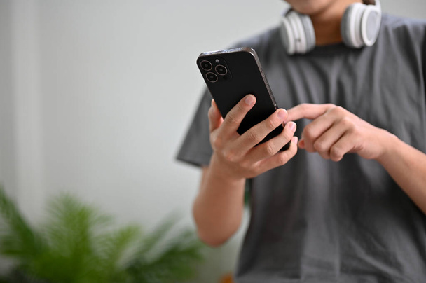 Oturma odasında günlük giysiler içinde genç bir Asyalı arkadaşlarıyla mesajlaşmak için akıllı telefonu kullanıyor, yeni başvuru formunu test ediyor, mobil uygulama üzerinden yemek sipariş ediyor. kırpılmış resim - Fotoğraf, Görsel