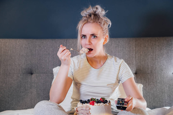 Emotionally stressed, shocked woman eating cake and holding a remote control while watching TV in bed. Scary horror movie or terrible news, scare in her eyes Overeating, Jamming of negative emotions - Φωτογραφία, εικόνα