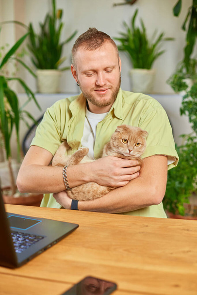 Man is working on a black notebook, laptop and cat is laying in his hand, Work from home, Freelancer remote working at home, green house, workplace at home. - Photo, Image