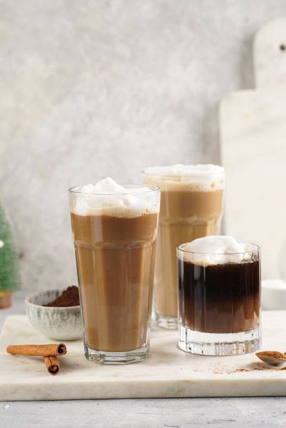 Three tall glasses with warm coffee drink with cinnamon, whipped milk foam and caramel on a marble board and christmas ornaments and decoration - Fotografie, Obrázek