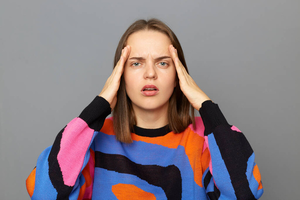 indoor shot of sad unhappy shocked woman wearing colorful sweater posing isolated over gray background, keeps hands on her temples, being frustrated, suffering headache, has problems. - Фото, зображення