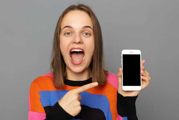 Portrait of happy amazed woman with brown hair wearing colorful jumper, pointing at smart phone with smart phone with empty display, keeps mouth open, posing isolated over gray background. - Фото, изображение