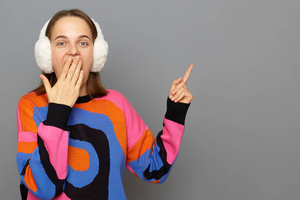 Portrait of surprised astonished woman wearing colorful sweater posing isolated over gray background, covering mouth with palm, pointing away at advertisement area. - Фото, зображення