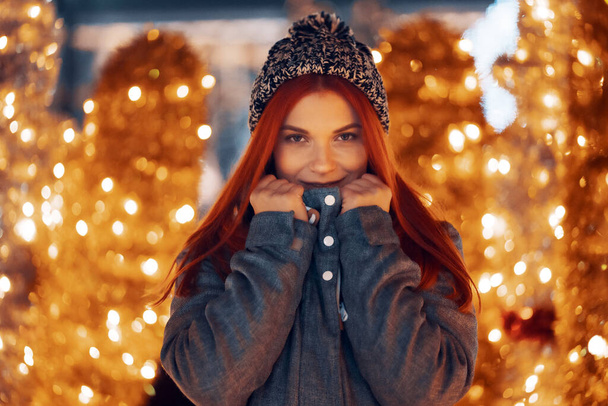 Outdoor night photo of young beautiful happy smiling girl enjoying festive decoration, posing in Christmas fair, in street of european city, wearing knitted beanie hat - Photo, Image