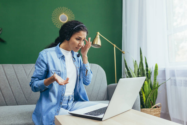 Serious young hispanic woman working from home, consulting, help desk, customer service. Sitting on the sofa with a laptop and headphones on a video call. - 写真・画像