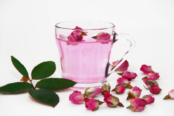 té rosa en una taza de vidrio transparente y pequeños brotes de rosa rosa seca sobre un fondo blanco - Foto, imagen