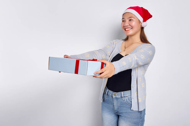 サンタクロースの帽子をかぶった青いセーターを着た笑顔の美しい若いアジアの女性の肖像画は、白いスタジオの背景に孤立したギフトボックスを与えます。メリークリスマスコンセプト - 写真・画像