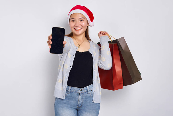 Joven mujer asiática alegre usando un sombrero de Navidad llevando bolsas de compras y mostrando las promociones de Navidad recomendadas en el teléfono móvil aislado sobre fondo blanco - Foto, imagen