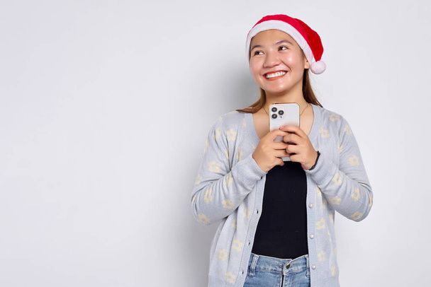 Χαμογελώντας νεαρή Ασιάτισσα με ένα χριστουγεννιάτικο καπέλο κρατώντας το κινητό τηλέφωνο και κοιτάζοντας κατά μέρος το χώρο αντίγραφο απομονώνονται σε λευκό φόντο - Φωτογραφία, εικόνα