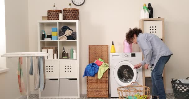 Um adolescente sorridente de jeans e uma camisa se senta na frente de uma máquina de lavar roupa caseira. Ele carrega roupas sujas na máquina de lavar roupa. Uma lavandaria brilhante e arejada. - Filmagem, Vídeo