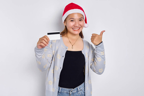 Sonriente joven asiática en un sombrero de Navidad que muestra la tarjeta de crédito blanca en blanco y el gesto de pulgar hacia arriba aislado en el fondo blanco - Foto, imagen
