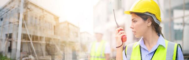 Jovem mulher de construção atraente em colete de segurança com capacete amarelo trabalhando com rádio, de pé no canteiro de obras de construção. Projecto de construção de casa - Foto, Imagem