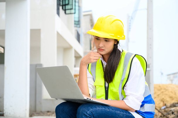 Giovane donna da costruzione attraente sorridente con casco giallo che lavora con il computer portatile, seduto in cantiere. Progetto di costruzione casa - Foto, immagini